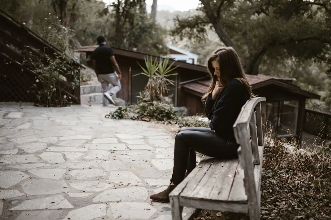 A woman sits pensively on a bench outside after a breakup, while a man walks away.